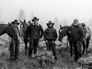 A black and white shot of stoney-faced ( Quarter Circle Ranch hands, wranglers, and cowboys staring straight into the camera. They are featured with their horses.