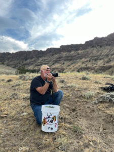 The photographer featured, camera in hand, with a bucket to sit on for stabilization while shooting the landscape.